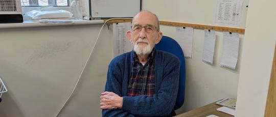 Image of elderly man sitting at his desk with laptop and papers looking at camera
